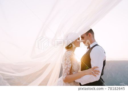 Fine-art destination wedding photo in Montenegro, Mount Lovchen. The wedding couple hugs on top of the mountain at sunset, the groom hugs the bride by the waist, the veil waved into the air. Fine-art destination wedding photo in Montenegro, Mount Lovchen. The wedding couple hugs on top of the mountain at sunset, the groom hugs the bride by the waist, the veil waved into the air. 78182645