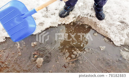 Close-up of a child's feet in rubber boots, a boy child happily playing with a shovel in a muddy puddle with snow and ice, in the early spring in the yard Close-up of a child's feet in rubber boots, a boy child happily playing with a shovel in a muddy puddle with snow and ice, in the early spring in the yard 78182883