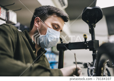 Bicycle shop repairman works in bicycle service and repair workshop during coronavirus quarantine wearing face shield and gloves, new norm. Mechanic repairing bicycle wheel wearing medical mask 78184107
