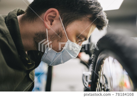 Bicycle mechanic wearing protective mask and gloves repairs customers bicycle wheel in accordance with quarantine standards during coronavirus pandemic. Small business during covid 19 lockdown 78184108