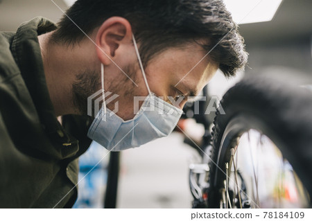 Mechanic repairs bicycle in workshop wearing covid 19 mask. Worker of bicycle store fixes bike during coronavirus quarantine in protective mask. Repairman maintenance cycle. Sport shop concept 78184109