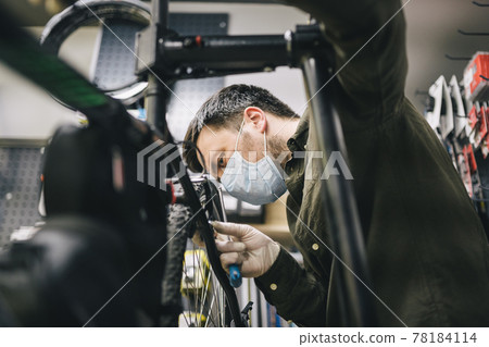 Bicycle mechanic wearing protective mask and gloves repairs customers bicycle wheel in accordance with quarantine standards during coronavirus pandemic. Small business during covid 19 lockdown 78184114