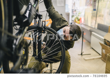 Bicycle mechanic wearing protective mask and gloves repairs customers bicycle wheel in accordance with quarantine standards during coronavirus pandemic. Small business during covid 19 lockdown 78184695