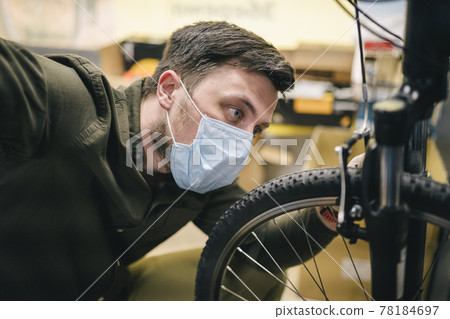 Bicycle shop repairman works in bicycle service and repair workshop during coronavirus quarantine wearing face shield and gloves, new norm. Mechanic repairing bicycle wheel wearing medical mask 78184697