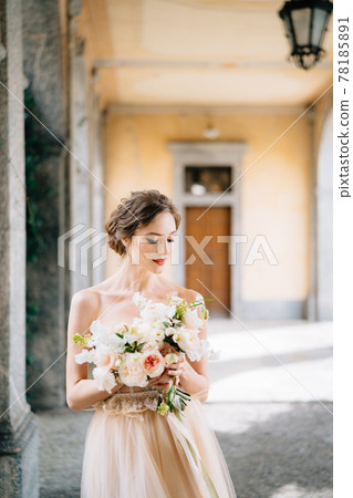 Bride in a beautiful dress with a bouquet of pink flowers stands in an arched corridor. Lake Como, Italy Bride in a beautiful dress with a bouquet of pink flowers stands in an arched corridor. Lake Como, Italy 78185891