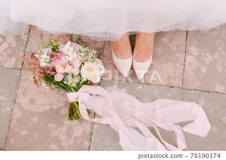 The bride's legs in graceful white shoes peek out from under the skirt, next to the bride's bouquet lies, close-up The bride's legs in graceful white shoes peek out from under the skirt, next to the bride's bouquet lies, close-up 78190174