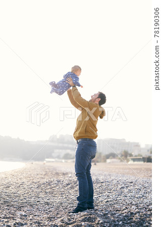 Smiling dad in a brown jacket and jeans throws a small child in a blue overalls high on his arms while standing on a pebble beach Smiling dad in a brown jacket and jeans throws a small child in a blue overalls high on his arms while standing on a pebble beach 78190306