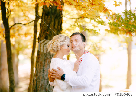 Half-length portrait of a man and a woman in white sweaters hugging against the background of trees in the autumn forest Half-length portrait of a man and a woman in white sweaters hugging against the background of trees in the autumn forest 78190403