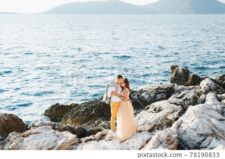 Handsome groom hugs cute smiling bride in pastel dress while standing on cliff above sea 78190833