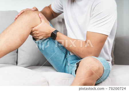 The man suffering from knee pain sitting on the couch hold and massaging his painful knee. The man suffering from knee pain sitting on the couch hold and massaging his painful knee. 78200724
