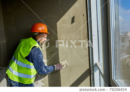Worker in workwear and safety helmet is putting putty on wall, using spatula. Apartment of building under construction with panoramic windows 78206934