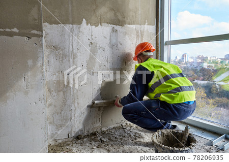 Skilled worker in working clothes and orange hard hat is putting putty on the wall, using spatula. Flat of a building under construction 78206935