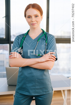 Portrait of confident female doctor in blue green uniform standing on background of window in sunny day in medical clinic office. Portrait of confident female doctor in blue green uniform standing on background of window in sunny day in medical clinic office. 78207402