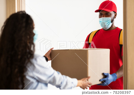 Black delivery man in mask giving cardboard box to woman 78208397