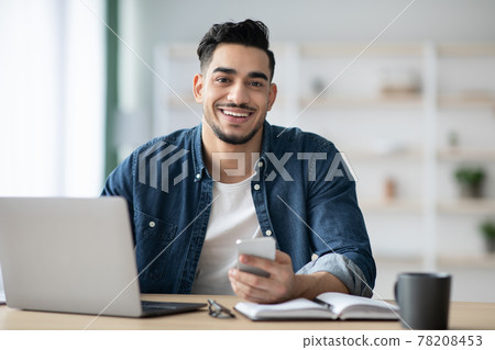 Smiling arab guy using smartphone while sitting at workdesk Smiling arab guy using smartphone while sitting at workdesk 78208453
