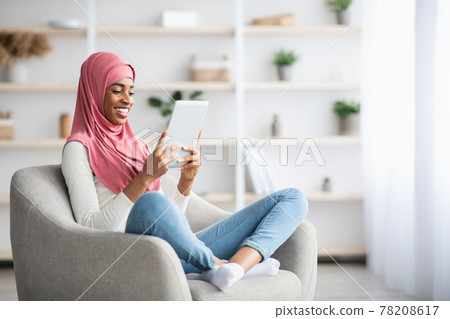 Young Black Muslim Woman In Hijab Relaxing In Chair With Digital Tablet Young Black Muslim Woman In Hijab Relaxing In Chair With Digital Tablet 78208617