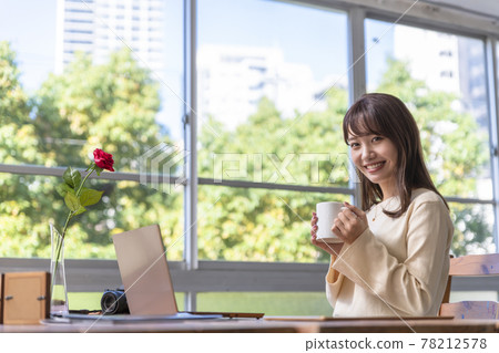 A woman in her twenties sitting in front of a computer by the window and drinking coffee A woman in her twenties sitting in front of a computer by the window and drinking coffee 78212578