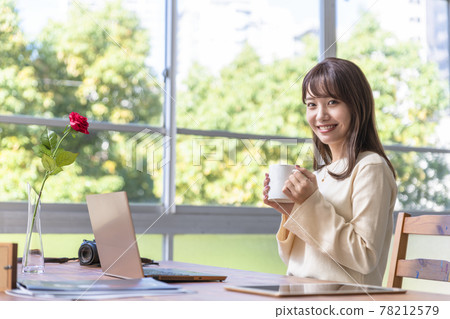 A woman in her twenties sitting in front of a computer by the window and drinking coffee 78212579