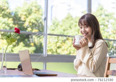 A woman in her twenties sitting in front of a computer by the window and drinking coffee A woman in her twenties sitting in front of a computer by the window and drinking coffee 78212580