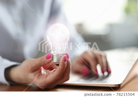 Woman holds light bulb in her hand and works on laptop 78215504