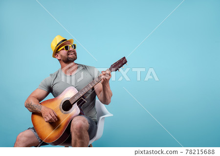 Handsome bearded guitarist in yellow hat and sunglasses sitting on chair and singing while playing on acoustic guitar isolated on blue background 78215688