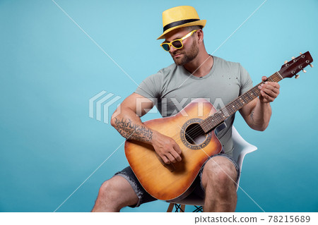 Handsome bearded guitarist in yellow hat and sunglasses sitting on chair and singing while playing on acoustic guitar isolated on blue background 78215689