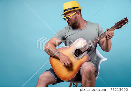 Handsome bearded guitarist in yellow hat and sunglasses sitting on chair and singing while playing on acoustic guitar isolated on blue background Handsome bearded guitarist in yellow hat and sunglasses sitting on chair and singing while playing on acoustic guitar isolated on blue background 78215690