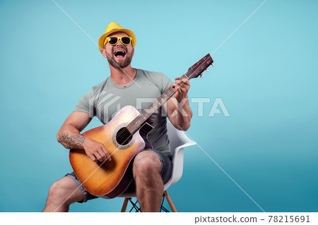 Handsome bearded guitarist in yellow hat and sunglasses sitting on chair and singing while playing on acoustic guitar isolated on blue background Handsome bearded guitarist in yellow hat and sunglasses sitting on chair and singing while playing on acoustic guitar isolated on blue background 78215691