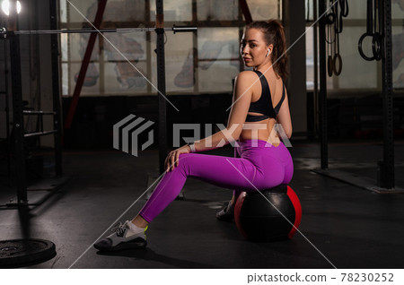 Beautiful young sporty woman is resting sitting on a medicine ball after active training in the gym. The girl listens to music on wireless headphones during fitness. Circular training. 78230252