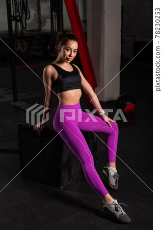 Beautiful young caucasian girl posing in a dark gym. A woman in sportswear is resting from a circular workout on a box. A break between sets. Beautiful young caucasian girl posing in a dark gym. A woman in sportswear is resting from a circular workout on a box. A break between sets. 78230253