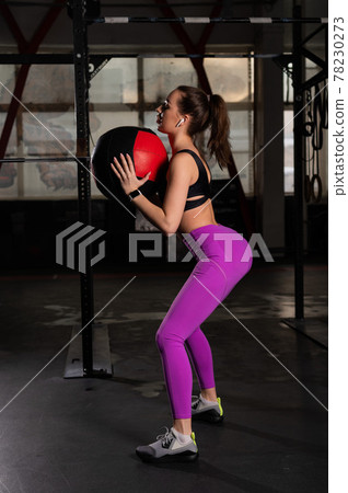 Beautiful European woman throws a cross fit ball. A girl in sportswear listens to music on a wireless headphone during a circle workout. Brunette is jumping. 78230273