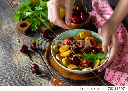 In the hands of a woman breakfast. Mini pancakes with cherries and honey on a rustic wooden table. In the hands of a woman breakfast. Mini pancakes with cherries and honey on a rustic wooden table. 78231297