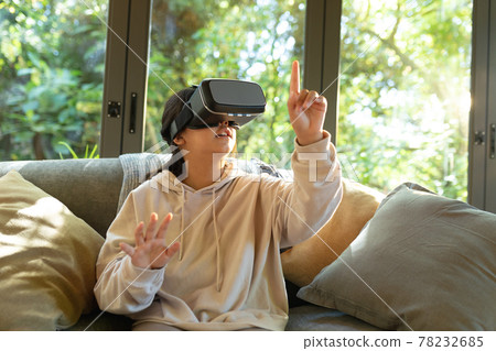 Smiling asian girl wearing vr headset touching with her finger sitting on sofa Smiling asian girl wearing vr headset touching with her finger sitting on sofa 78232685