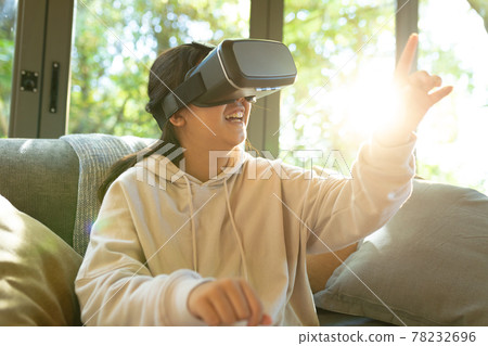 Smiling asian girl wearing vr headset touching with her finger sitting on sofa Smiling asian girl wearing vr headset touching with her finger sitting on sofa 78232696