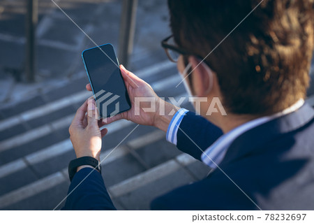 Asian businessman wearing face mask using smartphone sitting on steps in city street Asian businessman wearing face mask using smartphone sitting on steps in city street 78232697