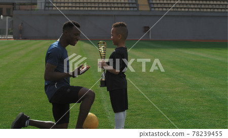 Black trainer giving cup to boy on football pitch 78239455