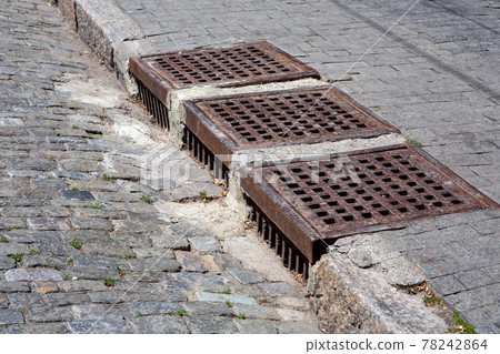 drainage system of urban infrastructure with an iron grating of the manhole grille of the stormwater system of the sidewalk from granite tiles near road from the paving stones on sunny day, nobody. 78242864