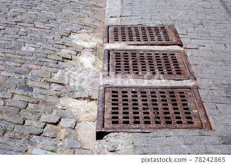 drainage system of city infrastructure with rusty iron grating of manhole grille of the stormwater system of sidewalk from granite tiles near road from paving cobblestones on sunny day, nobody. 78242865