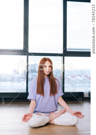 Serene attractive young woman meditating sitting on floor in lotus pose on background of window in light office room. Serene attractive young woman meditating sitting on floor in lotus pose on background of window in light office room. 78245213