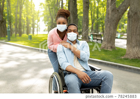 Millennial black woman hugging her handicapped boyfriend in wheelchair, wearing face masks, going for walk at city park Millennial black woman hugging her handicapped boyfriend in wheelchair, wearing face masks, going for walk at city park 78245883