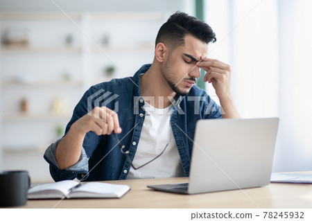 Exhausted arab guy sitting at workdesk, removing glasses Exhausted arab guy sitting at workdesk, removing glasses 78245932