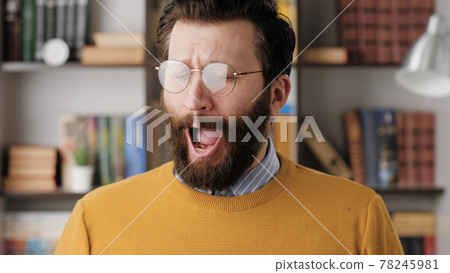 Man yawns. Tired sad bearded man in glasses in office or apartment room yawns, he is bored or wants to sleep. Close up Man yawns. Tired sad bearded man in glasses in office or apartment room yawns, he is bored or wants to sleep. Close up 78245981