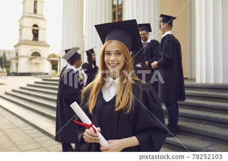 Young smiling girl university graduate in traditional mantle standing and holding diploma in hand Young smiling girl university graduate in traditional mantle standing and holding diploma in hand 78252103