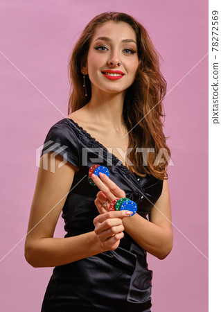 Brunette model with earring in nose, in black dress. She smiling, holding two blue and red chips, posing on pink background. Poker, casino. Close-up 78272569