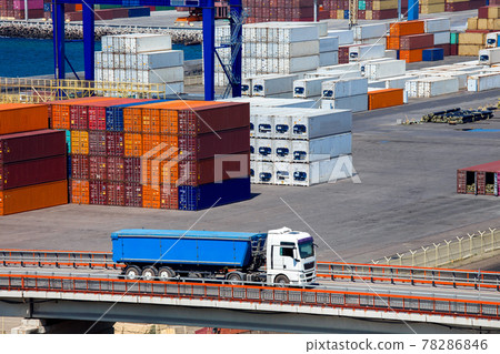truck with a trailer rides over a bridge in the background a warehouse of a merchant port with stacked containers of export goods brought by a ship along the sea route. truck with a trailer rides over a bridge in the background a warehouse of a merchant port with stacked containers of export goods brought by a ship along the sea route. 78286846