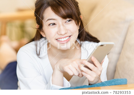A woman lying on the sofa in the living room and looking at the screen of her smartphone 78293353