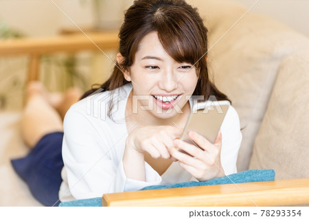 A woman lying on the sofa in the living room and looking at the screen of her smartphone 78293354