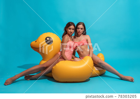 Two girls in swimsuits posing on inflatable duck on blue background Two girls in swimsuits posing on inflatable duck on blue background 78303205
