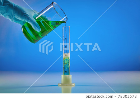 Hand in disposable glove is pouring green liquid from beaker into measuring cylinder, blue background. Coronavirus. Worldwide pandemic COVID-19 78303578