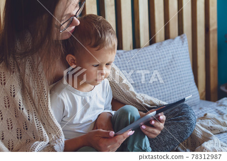 Young mom and son with a tablet under a cozy blanket in the bedr 78312987